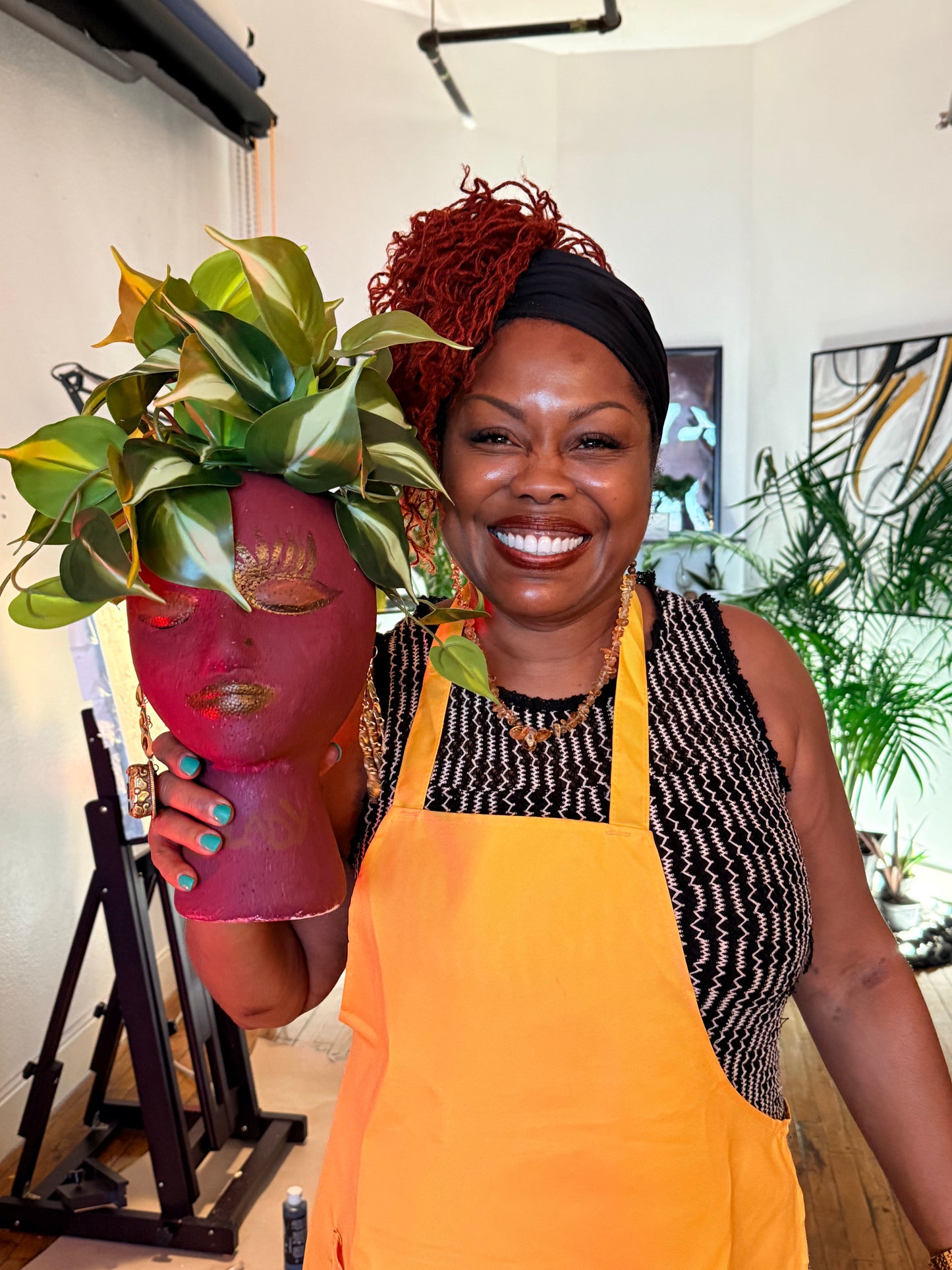 Woman wearing an apron holding a pink head sculpture with green leaves, in a room with plants and art.