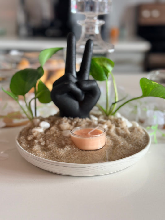 Black hand making a peace sign on a sand-like surface with plants and a candle.